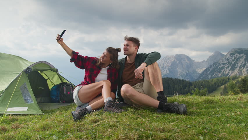 Cheerful couple having fun by their tent on a mountain camping spot, taking memorable selfie photos with a smartphone