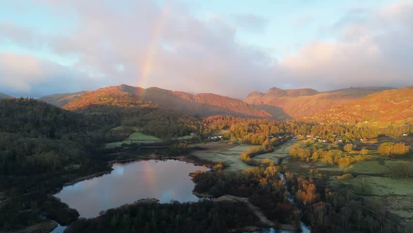 An aerial view showcases a stunning rainbow arching over a picturesque landscape of mountains, a lake, and lush forests under a dramatic sky.