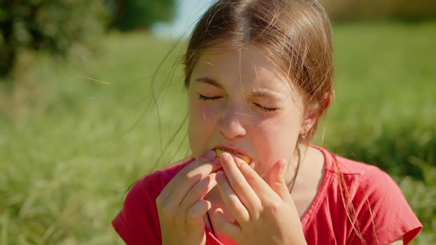 Young girl eating sour lemon, making expressive face. Kid tasting tart lemon outdoors, reacting strongly to sourness. Child biting into lemon slice, showing exaggerated sour reaction in sunny