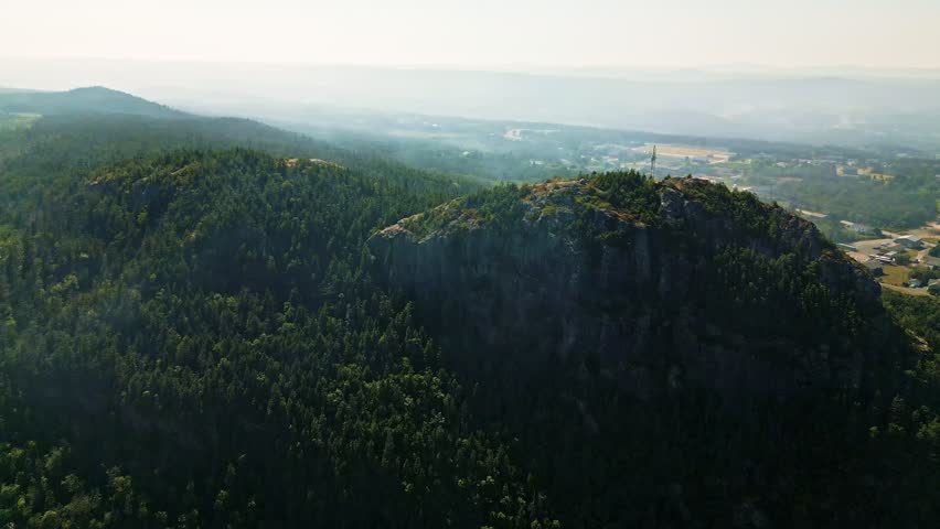 A drone glides over Holyrood's dark green hills and valleys while a generating station's smoke plume drifts across the sky, blending with the hazy horizon.