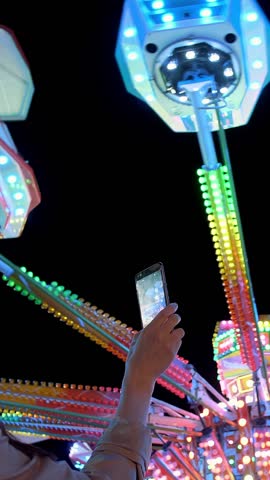 Woman hands are holding smartphone and taking photo or shooting video. Carousel with lights. Incredible colorful flashing light of vintage carousel. Popular Chair Swing Rides in Amusement Park