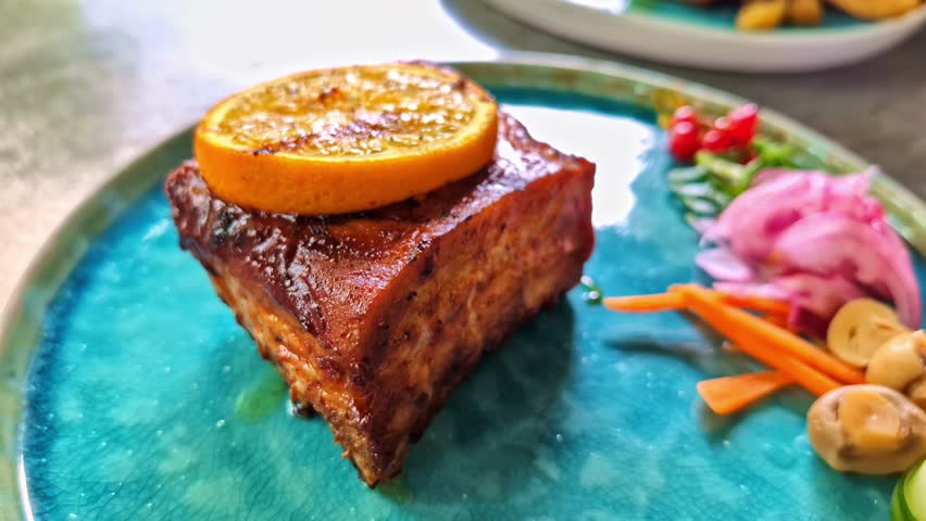 Slow parallax effect reveals a tender glazed pork belly topped with orange slice, artful pickled vegetables, and mushrooms on a blue ceramic plate.