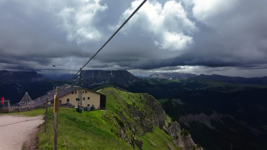 Scenic view from Seceda peak in the Dolomites with dramatic clouds, green cliffs, mountain hut, and flock of birds soaring over the ridge, Northern Italy