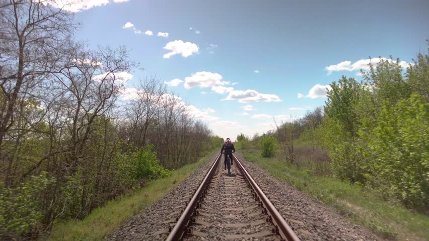 Male athletic cyclist riding bicycle on railroad track during summer trip. Cyclist riding along railway tracks under blue sky, exploring nature on adventurous bike tour.