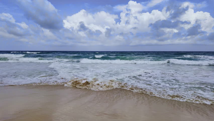 A dramatic view of powerful ocean waves crashing onto a sandy beach under a cloudy, stormy sky. The scene captures the raw energy of nature and the dynamic motion of the sea.