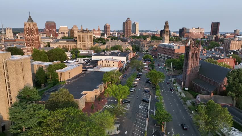 Aerial view of downtown New Haven, Connecticut, showing historic churches, Yale University buildings and modern skyscrapers in warm evening light. Waving American flag in proud city. Wide shot.