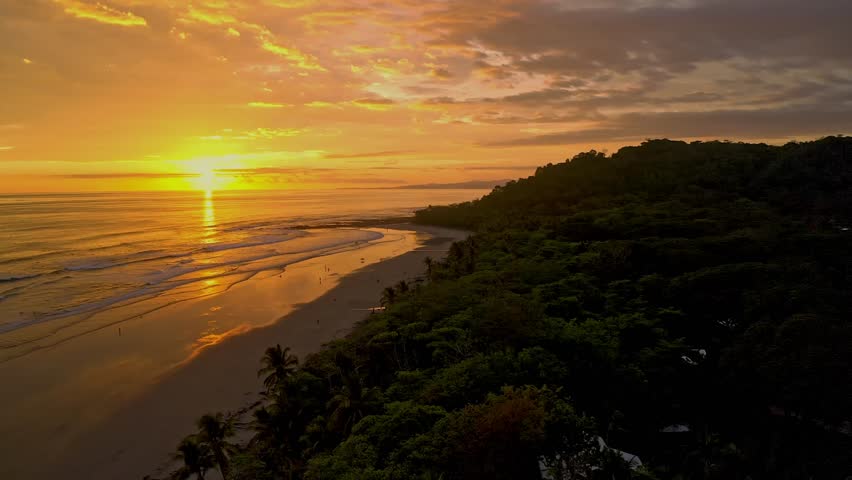 An aerial view captures the breathtaking sunset over Santa Teresa Coast in Costa Rica, with golden waves lapping the beach and lush hills framing the scene.