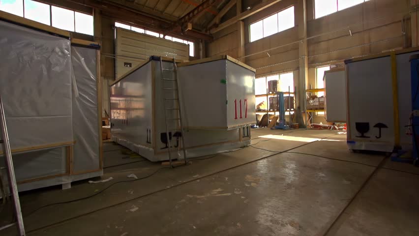 Empty prefab housing units rest inside open air warehouse with overhead lights and beams