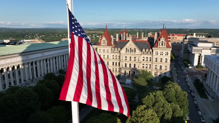 Waving American flag on flagpole at west capitol park in Albany. Aerial closeup. Chancellor hall and New York State capitol building in distance. Golden hour in the evening. Summer season.