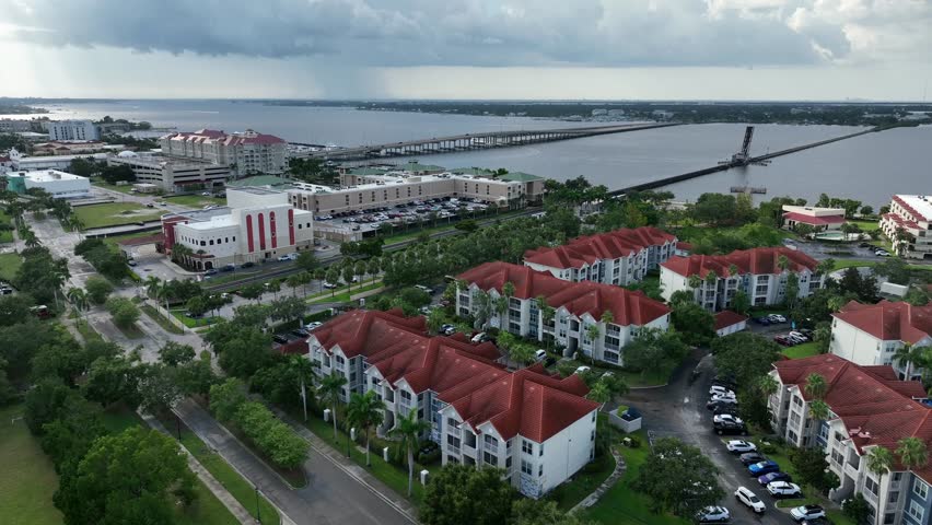 Aerial flyover Bradenton, Florida, showing red-roof residential complexes, waterfront buildings and long bridge spanning across the bay under cloudy day.