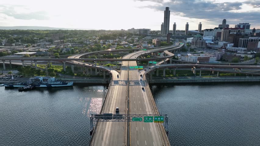 Driving cars on Dunn Memorial Bridge leading to downtown of Albany, New York. Peaceful golden sunset in American town. Clouds at sky. Aerial wide shot flyover. Elevated highway roads in USA.