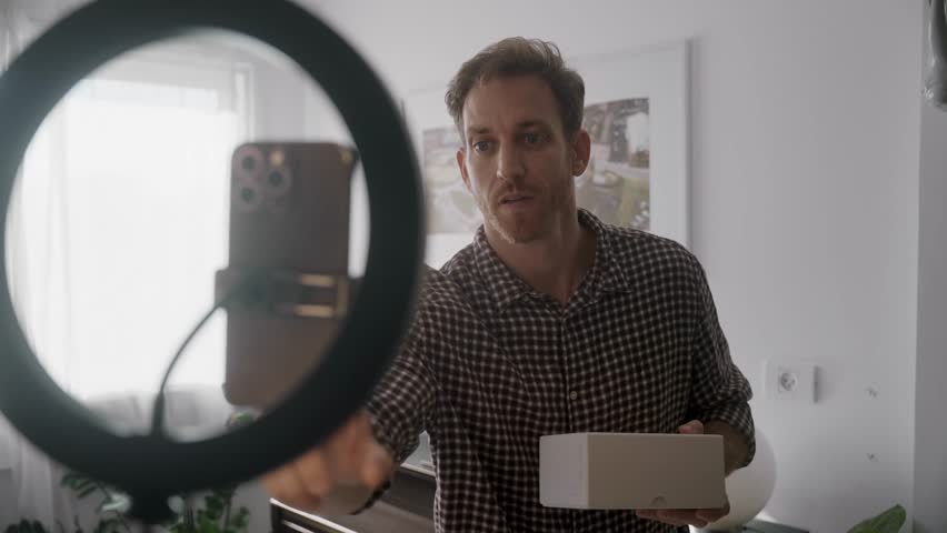 Man recording vlog or social media reel reviewing product unboxing video live stream. UGC style influencer talking to mobile phone in home studio. Excited and happy content creator opening white box