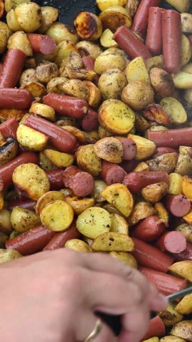 A person stirs seasoned young potatoes and cut sausages being fried in a large black pan over an open fire. Close-up view of the sizzling, hearty meal being prepared outdoors for a picnic. Vertical