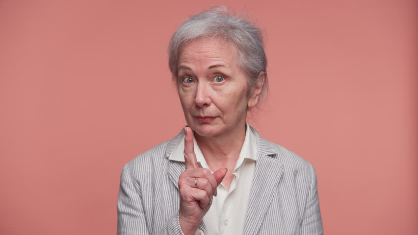 Serious senior woman giving warning gesture on pink background