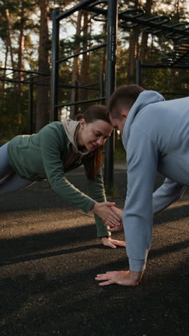 Vertical slow motion shot of young man and woman doing high five partner exercise while standing in plank in outdoor training area in forest