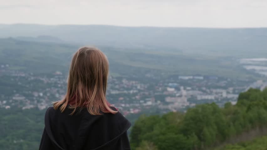 Blonde woman in black hoodie standing on hilltop, gazing across expansive valley with distant cityscape, embodying sense of freedom and peaceful solitude during outdoor exploration