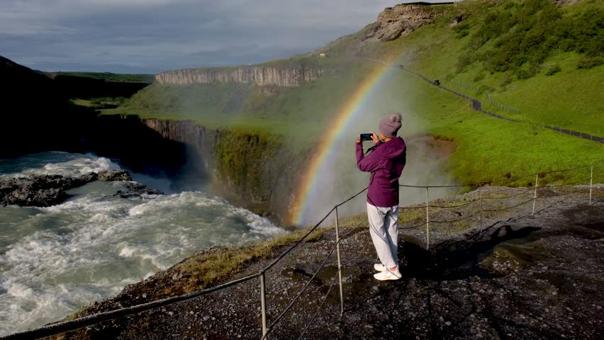 A traveler stands near Gullfoss waterfall, capturing the breathtaking sight of a vibrant rainbow arching over the raging water. Asian woman visiting Iceland on a trip