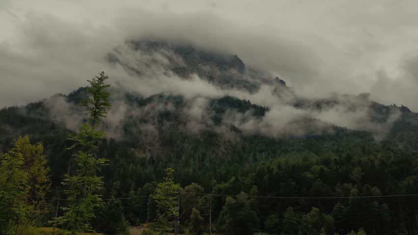 A wide panoramic view of the Austrian Alps wrapped in heavy clouds and rain. Dark stormy skies, misty atmosphere and dramatic mountains create a breathtaking mystical landscape.