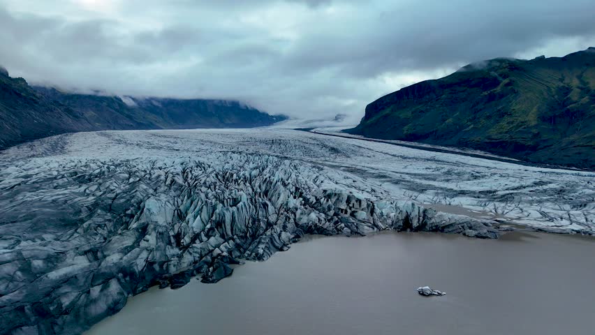 Explore the majestic Svinafellsjokull Glacier in Iceland, where intricate ice formations stretch across the landscape under a cloudy sky. The glacier flows towards distant peaks.