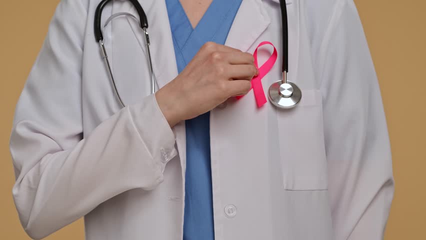 Doctor in white coat with stethoscope and pink ribbon on neutral background, points finger at ribbon as a symbol of awareness. The concept of breast cancer awareness day.
