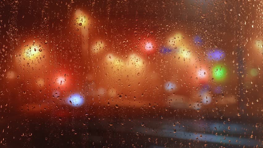 Raindrops dripping on a window pane with blurred multicolored city lights in the background at night. Bright bokeh effect creates a dreamy urban atmosphere.