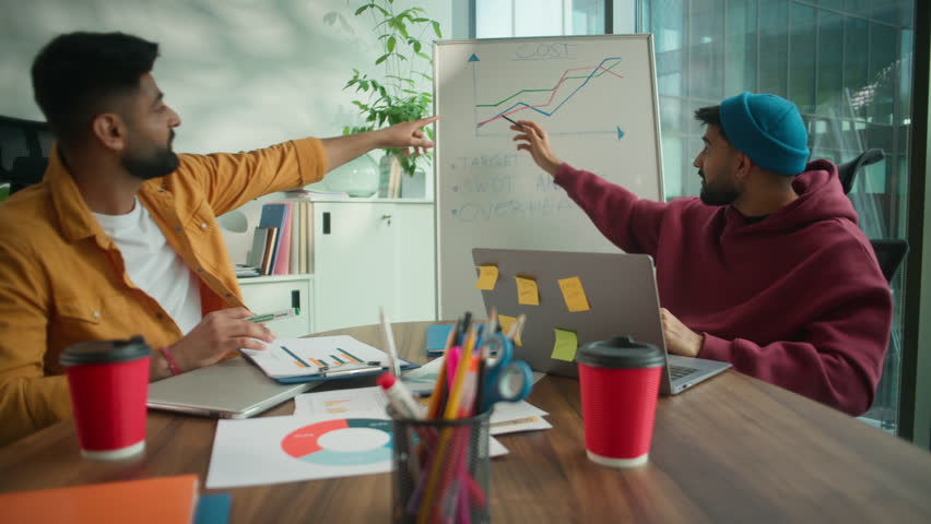 Two informal Indian businessmen Arabian business men colleagues males partners discussing strategy in modern coworking office pointing at whiteboard graph growth success thumbs up startup teamwork - Powered by Shutterstock - Get 15% off with code: PIKWIZARD15