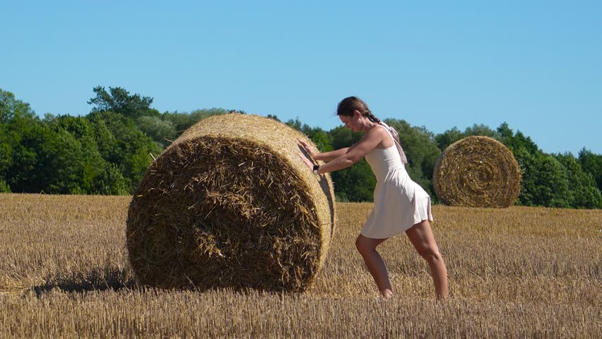 Field with bales. Woman pushing a bale of hay, woman in a summer field pushing hay gathered into a bale