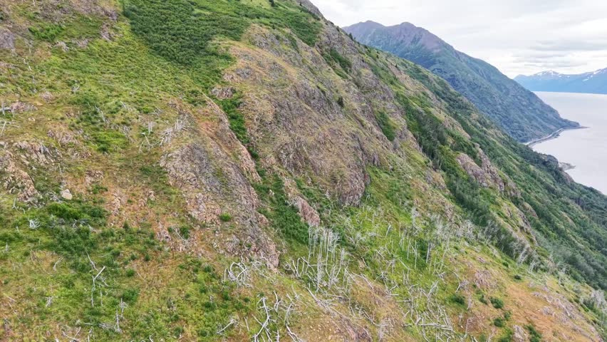 Drone aerial view of Beluga Point along the Seward Highway near Anchorage, Alaska. The image features dramatic mountain ranges, a curving coastal road, a railway line, and the calm waters of Turnagain