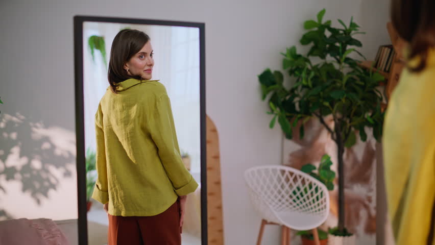 Stylish lady enjoying reflection trying elegant shirt in apartment. Smiling young woman looking mirror trying fashionable outfit at home. Beautiful brunette turning admiring appearance in living room.