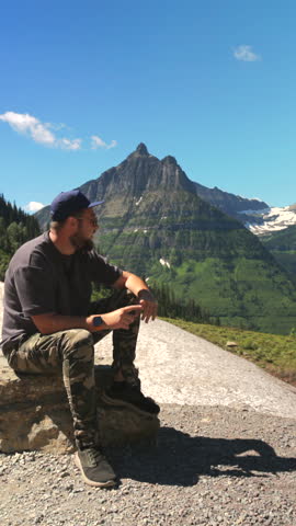 Man sitting facing the camera at Glacier National Park, showcasing stunning mountains and lush green landscapes. Its perfect for nature lovers and adventurers seeking unforgettable outdoor experiences