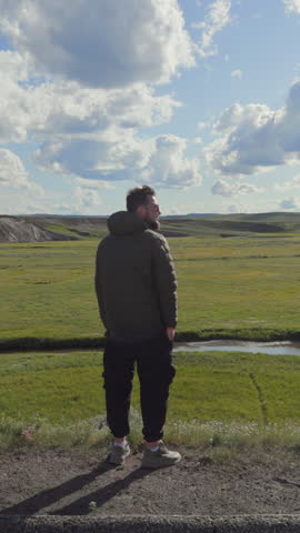 Male tourist stands at a scenic overlook at lamar valley at Yellowstone, gazing at the lush green fields and serene waters below, under a clear blue sky with fluffy clouds, beauty of nature