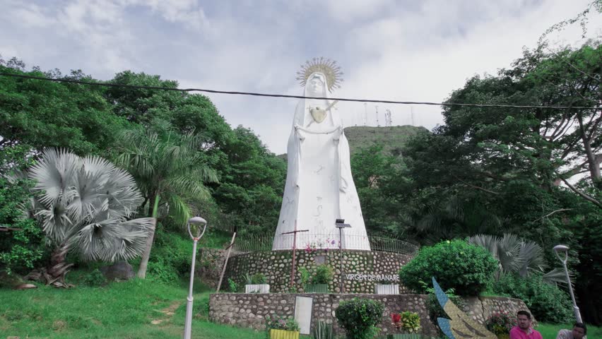 Drone zoom out showing the Virgen de Manare statue surrounded by lush vegetation in Yopal, Colombia, on a bright day.