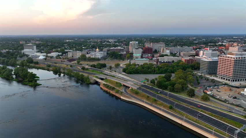 Downtown of Trenton with office buildings, cars on street along Delaware River. Golden sunset in New Jersey, bordering Pennsylvania state. Aerial backwards wide shot.