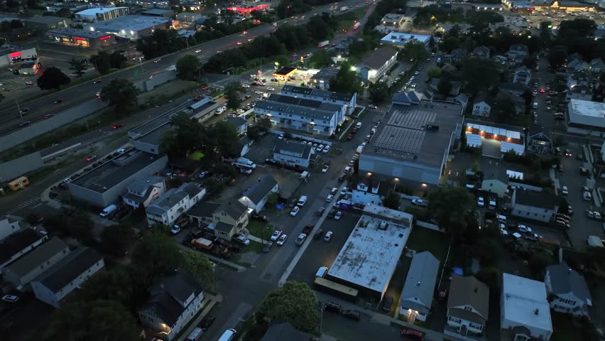 Stamford , United States - 08 03 2025: Aerial night flyover Stamford, Connecticut, with residential streets, commercial buildings and a highway with moving traffic under streetlights. Gas station and 