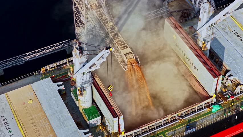A dynamic top-down aerial parallax shot shows a crane and chute loading grain into the open hold of a large bulk carrier ship at the industrial Port of Riga, Latvia.