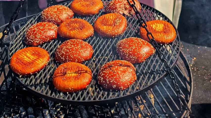A close-up shot shows sesame seed burger buns toasting on a hot grill over glowing charcoal embers at an outdoor street food market in Latvia, preparing for a delicious meal.