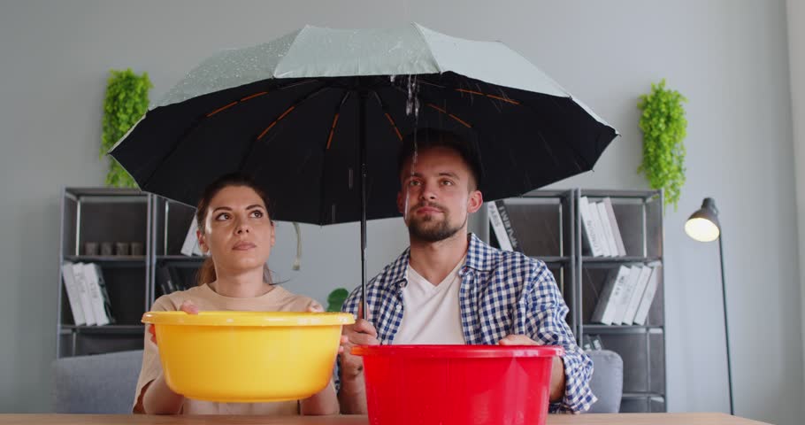 Frustrated couple sitting indoors under an umbrella with buckets collecting water from a leaking ceiling. Concept of housing issues, apartment maintenance, and home repair problems. 