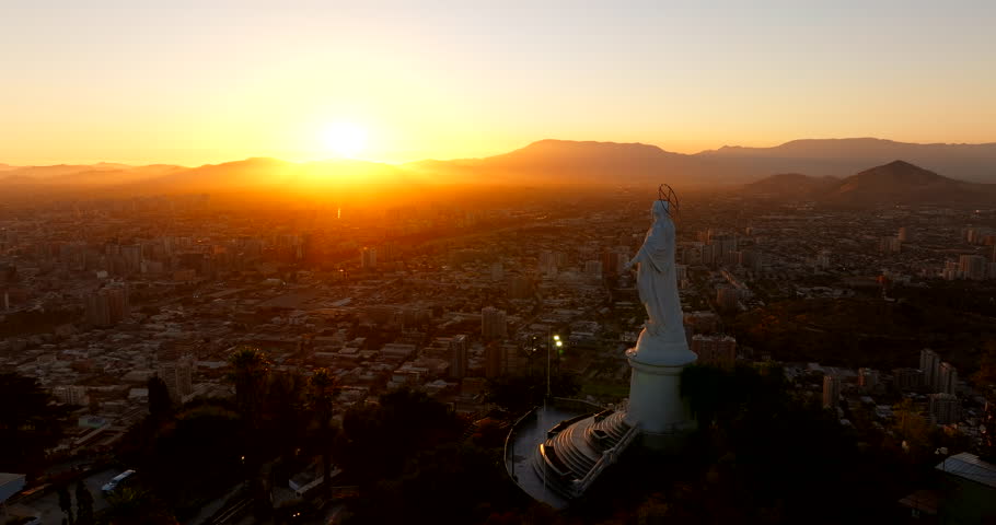 Forward-moving drone at sunset flies past the Sanctuary on San Cristóbal Hill, revealing the giant Immaculate Conception statue above Santiago and the Andes in warm golden haze.
