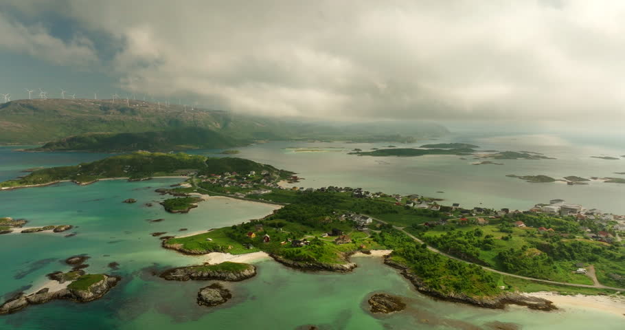 Drone pullback reveals Sommaroy islands and turquoise shallows near Tromsø, with a distant ridge-top wind farm and a broad fjord horizon under soft summer clouds.