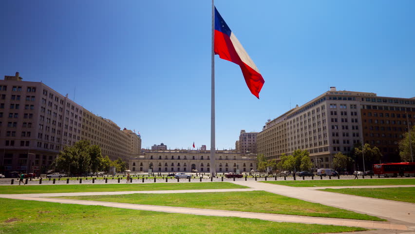 Chilean flag waves near the Centro Cultural La Moneda in downtown Santiago, Chile, towering above cars, static establishing