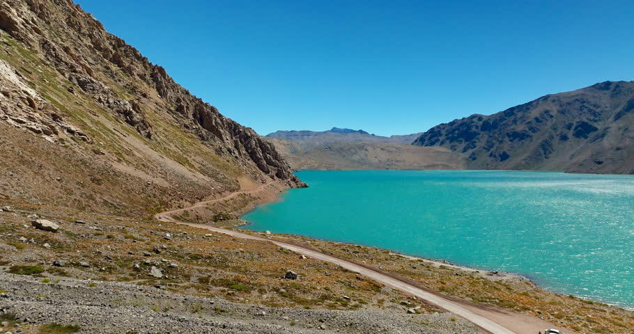 Low flight along a winding dirt road skirting a turquoise mountain lake in Cajon del Maipo, Chile, as the Andes rise on both sides of the canyon under clear summer skies.