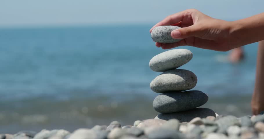 Female hands carefully placing pebbles on top of each other, creating a balanced stack on a pebble beach with the calm blue sea in the background
