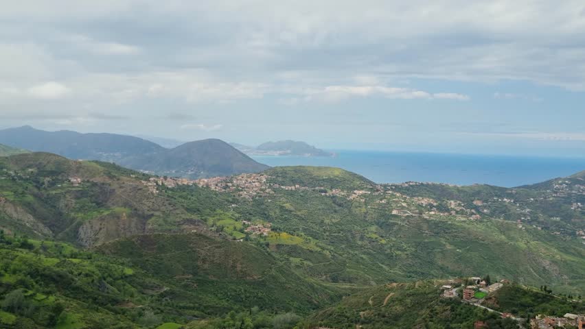 Shot from a mountain pass in the Tell Atlas region of Kabylie, Algeria, revealing green hills stretching to the Mediterranean Sea on a sunny day, capturing the vast, scenic landscape