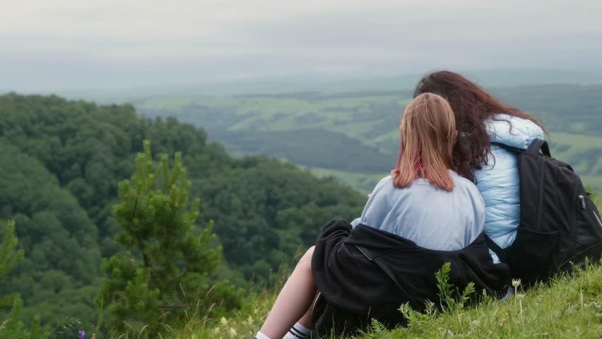 Female hikers resting on grassy mountaintop, gazing across expansive valley landscape, experiencing serene wilderness moment with breathtaking panoramic vista surrounding them