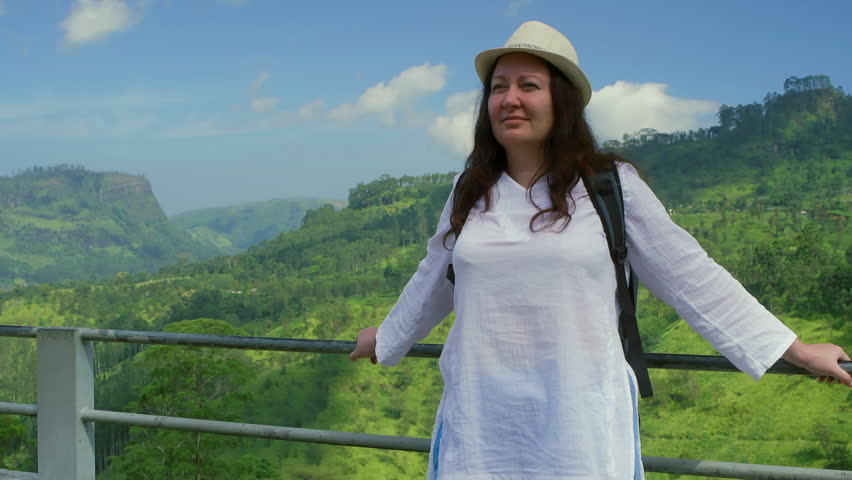 Female traveler in white dress and sunhat standing at railing, overlooking verdant tropical valley with lush green landscape under bright blue sky in Mauritius