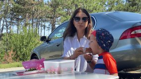 A woman is holding food, looking towards a child who is making a playful expression, as they sit at a portable table with a container of strawberries, all set beside a parked car on a sunny day - Powered by Shutterstock - Get 15% off with code: PIKWIZARD15