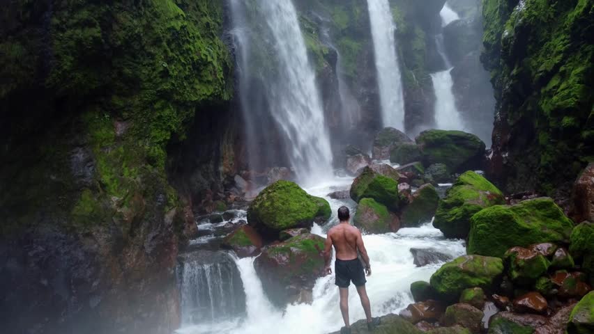 A breathtaking view of a man in the Quebrada Gata waterfalls in Costa Rica, showcasing multiple powerful cascades plunging into a rocky riverbed.
