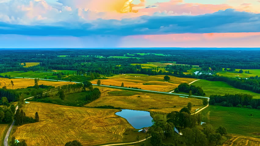 Golden glow obscured above storm clouds under daylight over green farmland and scattered farmhouses, reflection in water