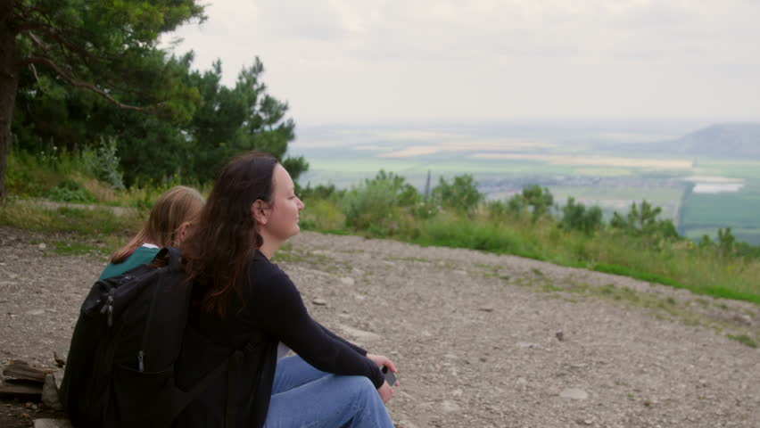 Mother and daughter hiking team resting at mountain summit, pausing during scenic journey and sharing peaceful moment while gazing across expansive green valley landscape under bright summer sky