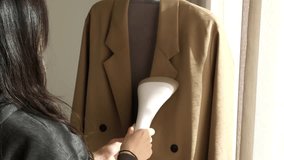 A woman using a handheld garment steamer to remove wrinkles from a brown jacket indoors. Concept of home laundry, fabric care, and daily household chores. - Powered by Shutterstock - Get 15% off with code: PIKWIZARD15